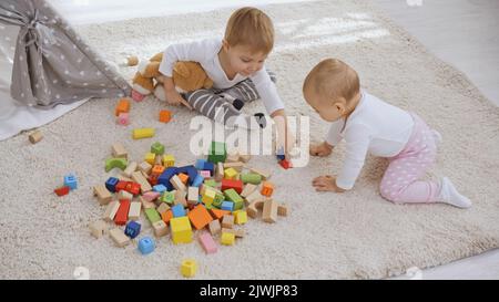 tout-petit garçon assis sur un tapis avec un ours en peluche près de sa sœur et jouant ensemble avec des blocs de bois Banque D'Images