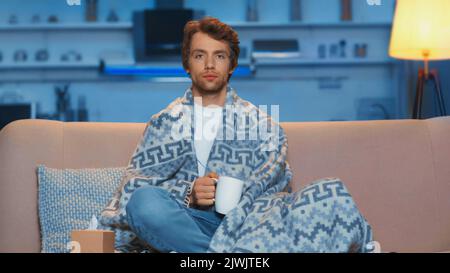 jeune homme malade assis sous une couverture et tenant une tasse de thé dans la salle de séjour Banque D'Images