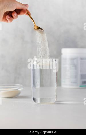 La poudre de collagène est versée dans un verre d'eau d'une cuillère dorée sur fond gris. Le concept de santé. Banque D'Images