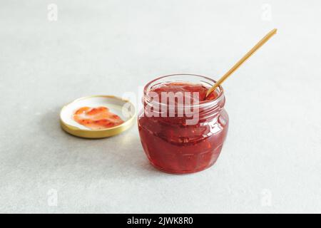 Dessert prêt à manger. Pot maison de confiture de fraises, cuillère dorée et couvercle sur fond gris. Style de vie vie encore vie, foyer doux sélectif. Banque D'Images