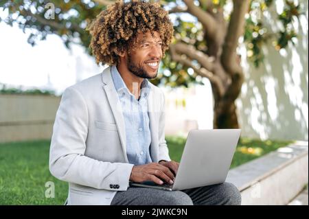 Calme pensif réussi latino positif ou brésilien homme d'affaires, développeur web, courtier, avec des cheveux bouclés, assis à l'extérieur avec un ordinateur portable, travaillant sur un projet à distance, regardant devant, sourires amicaux Banque D'Images