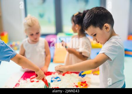 Détente sensorielle jeu avec sable cinétique moulable à l'école maternelle. Les tout-petits avec leur professeur s'amusent autour de la table en utilisant différents outils pour sculpter le sable, tels que des punaises texturées et colorées, des pinces coupantes, des étuis en silicone. Compétences motrices fines et développement créatif . Photo de haute qualité Banque D'Images