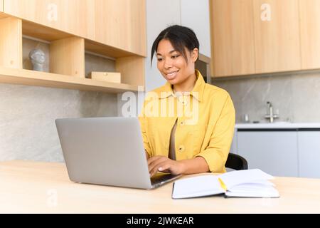 Portrait d'une jeune femme afro-américaine assise au bureau avec un ordinateur portable, en tapant un e-mail, en envoyant un message à un ami, en discutant, en datant en ligne avec un sourire agréable, concept de relations à distance Banque D'Images