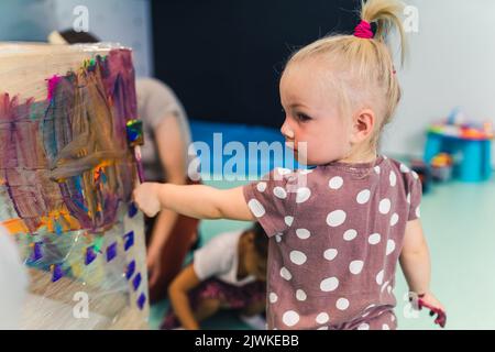 Peinture de film adhésif. Petite fille caucasienne enfant peinture avec ses bras sur un film adhésif enroulé tout autour de l'étagère en bois. Activité créative pour le développement des enfants à l'école maternelle. Photo de haute qualité Banque D'Images