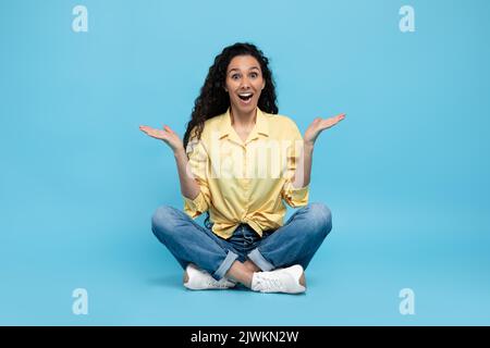 Une jeune femme excitée et perplexe se fait des épaules et souriant sur fond de studio bleu, sur toute la longueur Banque D'Images