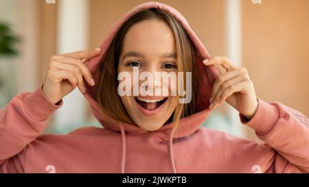 Portrait d'une jeune fille excitée portant une capuche et souriant à l'appareil photo, posant à l'intérieur à l'intérieur de la maison Banque D'Images