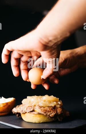 Main humaine ouvrant un œuf bouilli et le versant sur un hamburger Doble avec oignon cocké sur un fond noir Banque D'Images