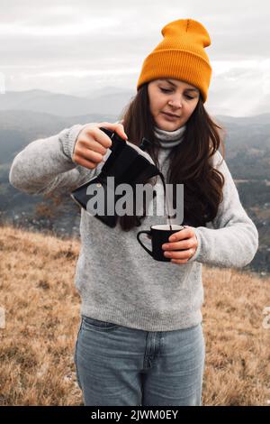 Femme en bonnet beanie orange qui verse le café de la marmite Moka dans une tasse à l'extérieur Banque D'Images