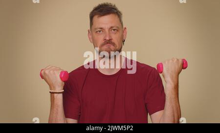 Beau jeune homme en t-shirt rouge s'entraîner, pompant les muscles du bras de levage rose gym haltères, pratiquant pilates, soins de santé. Homme de style adulte sur fond de mur beige studio. Motivation sportive Banque D'Images