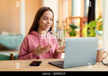 Une jeune fille positive visiophonie sur un ordinateur portable et gestuelle à la webcam, assis au bureau à la maison Banque D'Images