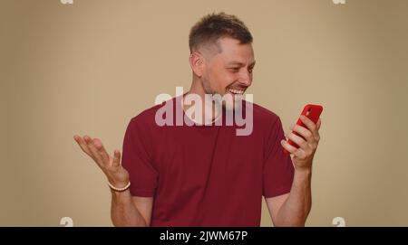 Joyeux joyeux joyeux jeune homme utiliser smartphone dactylographiant navigation dire wow oui, a découvert de grandes nouvelles de gain faisant le geste du gagnant. Homme de style adulte isolé sur fond de mur beige studio à l'intérieur Banque D'Images