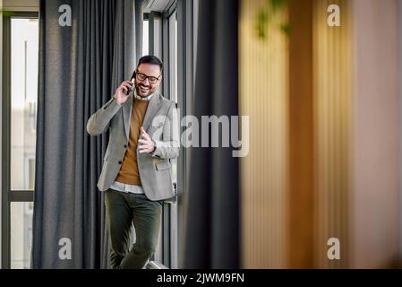 Homme adulte excité en costume, en train de raconter quelque chose de drôle à son collègue au téléphone, debout dans le hall. Banque D'Images