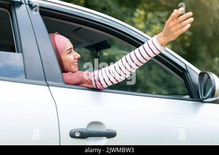 Bonne joyeuse jeune femme arabe dans hijab profiter de voyager sur la nouvelle voiture touche l'air avec sa main par la fenêtre ouverte Banque D'Images