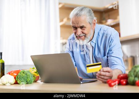 Homme âgé utilisant un ordinateur portable et une carte de crédit dans la cuisine, commander des articles d'épicerie en ligne Banque D'Images