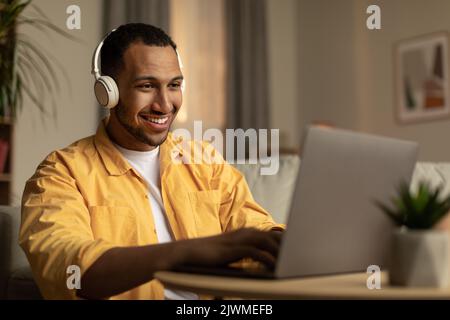 Joyeux jeune Africain américain utilisant un ordinateur portable, ayant un travail à distance, portant des écouteurs à la maison Banque D'Images