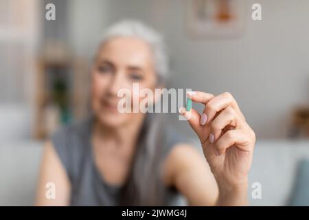 Gaie caucasienne femme plus âgée à cheveux gris tient la pilule dans la main pour le traitement de la maladie dans l'intérieur de la salle de séjour Banque D'Images