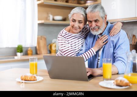 Un couple senior souriant utilise un ordinateur portable dans la cuisine tout en prenant le petit déjeuner ensemble Banque D'Images