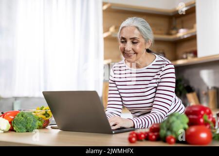 Portrait d'une dame âgée souriante utilisant un ordinateur portable dans la cuisine à la maison Banque D'Images