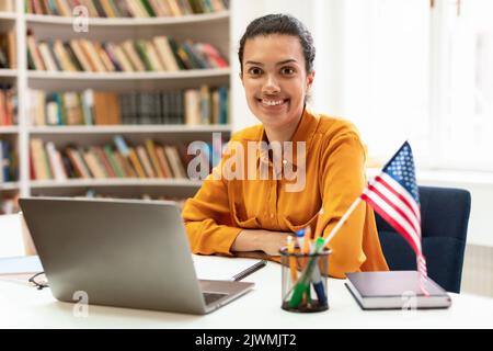Femme heureuse avec drapeau des Etats-Unis utilisant un ordinateur portable, étudiant l'anglais américain en ligne, assis dans la bibliothèque et souriant à la caméra Banque D'Images