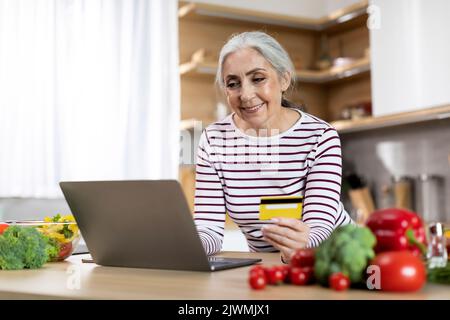 Femme âgée utilisant un ordinateur portable et carte de crédit dans la cuisine, commander des articles d'épicerie en ligne Banque D'Images