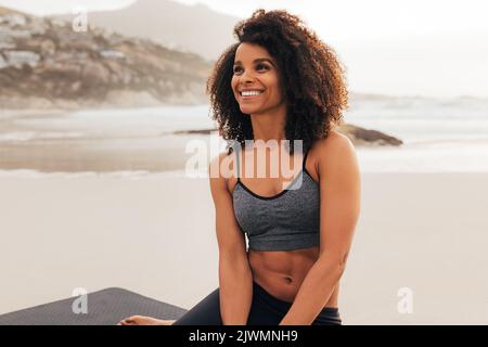 Portrait d'une belle jeune femme avec des cheveux bouclés portant des vêtements de sport assis sur la plage se détendant au coucher du soleil Banque D'Images