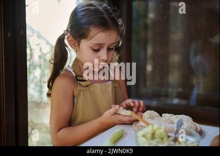 Magnifique petit cuisinier, adorable petite fille avec deux queues de cheval, dans les aliments du chef, écrasent les pommes de terre et les moules Banque D'Images