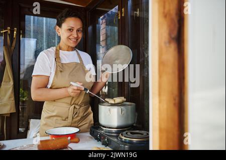 Femme charmante femme ménagère en tablier beige du chef, se tient près d'une cuisinière, fait des boulettes maison dans la casserole, prépare du varenyky Banque D'Images