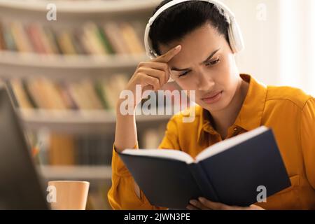 Enseignement à distance. Femme de race mixte concentrée dans un casque sans fil lisant le livre papier, assis à table dans la bibliothèque Banque D'Images