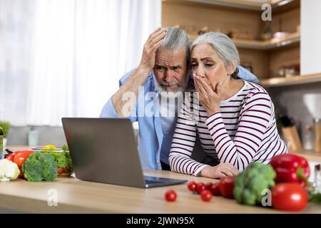 Un couple senior choqué regarde l'écran d'un ordinateur portable dans la cuisine Banque D'Images