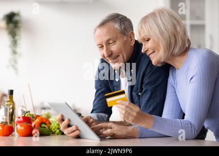 Couple senior utilisant une tablette numérique et une carte de crédit, cuisiner Banque D'Images