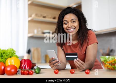 Jeune femme afro-américaine souriante avec smartphone et carte de crédit, shopping en ligne Banque D'Images