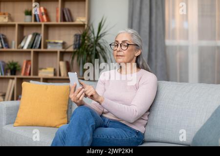 Femme grise, âgée, blanche et concentrée sérieuse, en lunettes de frappe sur le téléphone, utilisez une nouvelle application dans le salon Banque D'Images