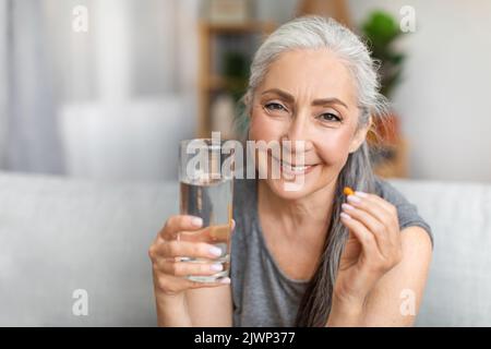 Une femme européenne aux cheveux gris très bonne et jolie affiche de la pilule et un verre d'eau dans le salon Banque D'Images