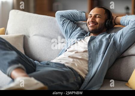 Souriant Africain américain Guy écouter de la musique tout en se relaxant sur le canapé à la maison Banque D'Images