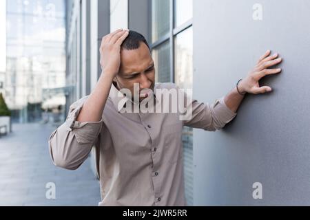Malade africain américain homme à l'extérieur du bâtiment moderne de bureau, l'homme a des maux de tête graves et de migraine, le travailleur en chemise sur la pause surfatigué. Banque D'Images