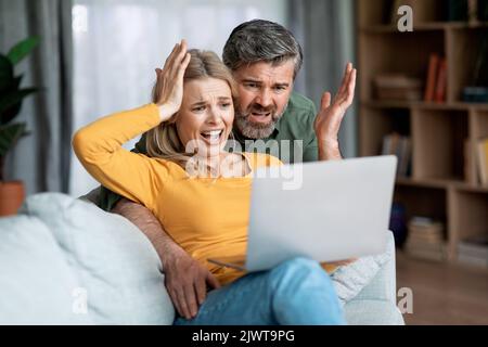 Concept de mauvaises nouvelles. Couple d'âge moyen choqué assis avec un ordinateur portable sur le canapé Banque D'Images