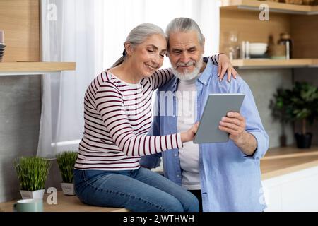 Les conjoints aînés heureux utilisant la tablette numérique tout en se relaxant dans la cuisine ensemble Banque D'Images