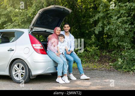 Reposez-vous et détendez-vous sur la route. Joyeux jeune arabe mâle et femelle dans le hijab avec un petit fils assis dans le coffre de voiture Banque D'Images