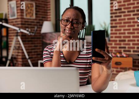 Une jeune femme afro-américaine prend son selfie sur son smartphone, souriant à l'avant de l'appareil photo. Travailleur indépendant qui fait des portraits sur un téléphone portable sur un lieu de travail à domicile, travailleur à distance qui parle lors d'un appel vidéo Banque D'Images