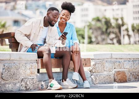 Couple noir, amour et téléphone tout en naviguant sur les médias sociaux, Internet ou en regardant une vidéo tout en étant assis sur un banc de parc. Homme et femme heureux sur un Banque D'Images