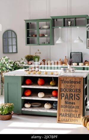 Cuisine avec mobilier vert vintage, comptoir en marbre avec fleurs et citrouilles, pots de fleurs d'automne, placard avec différentes tasses, vaisselle et cu Banque D'Images