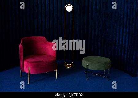 Un appartement avec un confortable fauteuil en velours rouge, un lampadaire minimaliste et un pouf vert dans une pièce avec des panneaux muraux en velours bleu foncé et le sol c Banque D'Images