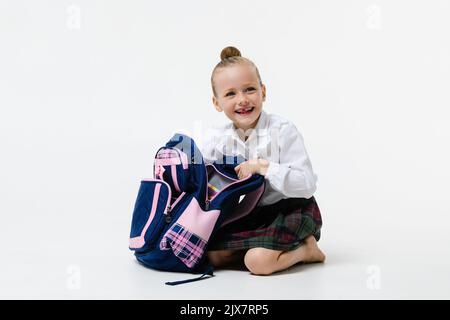 Mignonne fille en uniforme d'école ouvre un sac à dos pour les classes isolé sur un fond blanc. Banque D'Images