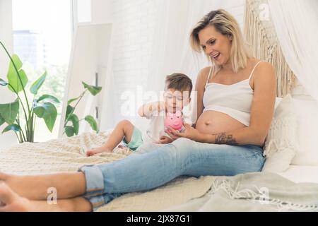 Photo en plein air d'une mère blonde millénaire caucasienne et de son tout-petit garçon de quatre ans assis ensemble sur un couvre-lit beige et mettant des pièces dans une jolie pigeonnaire rose. Concept d'économies. Photo de haute qualité Banque D'Images