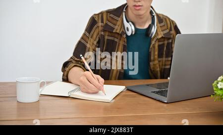 Un jeune enfant asiatique étudiant dans une chemise de flanelle travaillant sur son projet, recherche d'une information en ligne, en utilisant un ordinateur portable et en prenant des notes sur h Banque D'Images