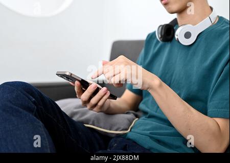 Homme asiatique millénaire assis sur un canapé et utilisant son smartphone. Image rognée et en gros plan. Banque D'Images