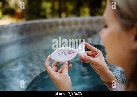 Jeune femme dans la baignoire, contrôle de la température de l'eau avec thermomètre, prêt pour la procédure de spa à la maison dans le bain à remous extérieur. Bien-être, soins du corps, hygiène Banque D'Images