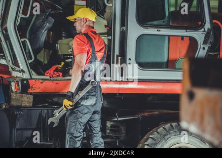 Mécanicien de machines à usage intensif debout devant la pelle hydraulique Orange avec de grandes clés entre ses mains. Atelier d'entretien et de réparation. Banque D'Images