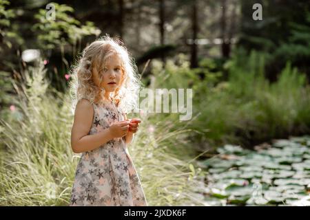 Portrait de la petite fille en pleurs bouleversés avec de longs cheveux bouclés moelleux debout près de l'étang avec des nénuphars verts dans le parc. Banque D'Images