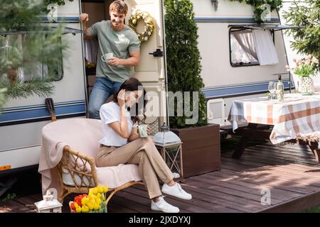 Homme souriant tenant une tasse près d'une petite amie sur un fauteuil près d'une camionnette de camping, image de stock Banque D'Images